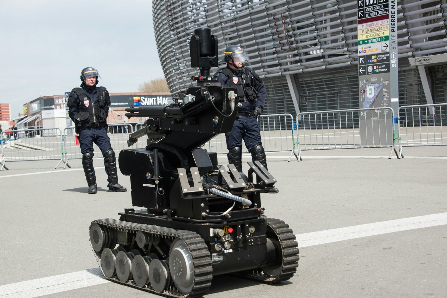 美国旧金山批准警察使用遥控武装机器人杀死嫌疑人 A picture of a black, tank-treaded robot outside a stadium flanked by two police officers in bomb protection gear.